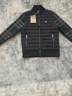Burberry Puffer Jacket - This is a Burberry puffer jacket in size Medium (M). The jacket features the classic Burberry check pattern on the sleeves and a quilted, padded body with zippered pockets. The inside label confirms the brand and size. The jacket is black with grey check detailing, and it has a front zipper closure. Vet inte varför det står på engelska, andra frågor skriv Dm
