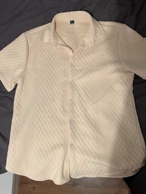Beige quiltad skjorta med kort ärm - Säljer en beige skjorta med kort ärm och quiltad struktur. Skjortan har klassisk krage, knappar framtill och ett unikt mönster i tyget som ger en cool textur. Perfekt för dig som vill sticka ut lite extra med din stil.