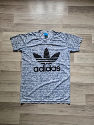 Blåmelerad Adidas t-shirt - Snygg blåmelerad t-shirt från Adidas med det klassiska svarta Trefoil-loggan tryckt på bröstet. T-shirten har rund halsringning och korta ärmar. Perfekt för träning eller chill. Materialet känns mjukt och lätt.