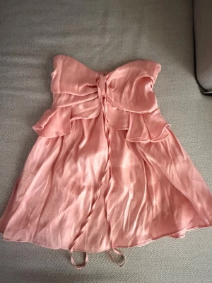 Ljusrosa axelbandslös tubtopp - Söt ljusrosa axelbandslös topp i ett glansigt material med volangdetaljer och knyt framtill. Perfekt för sommarens alla tillfällen. Klänningen har en feminin och flowy passform.