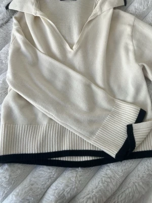 Vit stickad tröja med svarta detaljer - Klassisk vit stickad tröja med svarta ribbade kanter vid ärmslut, nederkant och krage. Tröjan har en djup v-ringning och bred krage som ger en avslappnad men stilren look. Perfekt till jeans eller kjol för en trendig vardagsstil.
