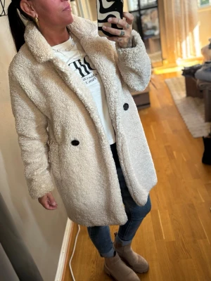 Bershka kappa - Mysig beige teddyjacka med dubbelknäppning och stora svarta knappar. Jackan har bred krage, två sidofickor och är tillverkad i fluffigt teddy-material. Perfekt för kalla dagar och ger en trendig look till din outfit. Créme vit