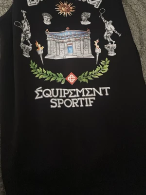 Svart hoodie med grafiskt tryck - Svart hoodie med stort grafiskt tryck på framsidan som visar statyer, byggnad, facklor och texten 'Équipement Sportif'. Hoodien har en klassisk passform och huva. Perfekt för dig som gillar unika och konstnärliga plagg.