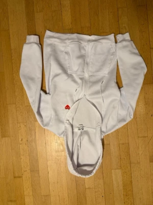 Vit hoodie med rött hjärta play - Säljer en vit hoodie med dragkedja och huva. På bröstet finns ett litet rött hjärta med ögon broderat. Hoodien har fickor framtill och snören i huvan. Perfekt för dig som gillar enkel men snygg streetwear.
