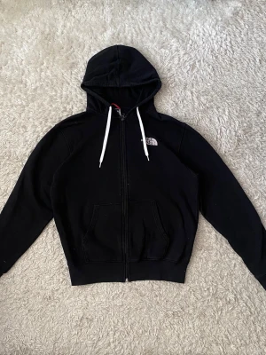 Svart hoodie från The North Face - Säljer en svart hoodie från The North Face med dragkedja, vita snören i huvan och logga broderad på bröstet. Hoodien har två fickor framtill och är tillverkad i mjukt material som passar perfekt för chill dagar.