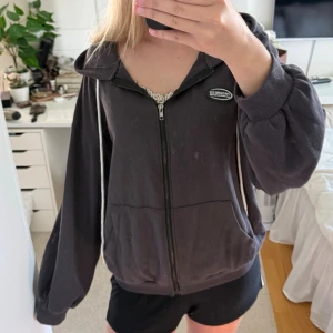 Svart zip hoodie med huva - En svart/grå hoodie med dragkedja framtill och huva med vita snören. Hoodien har en avslappnad passform, två fickor fram och ett litet märke på bröstet. Perfekt för en chill och casual look.