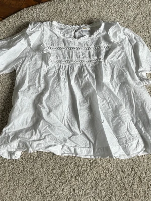 Vit blus med spetsdetaljer - Söt vit blus i luftig bomull med korta ärmar och fina spetsdetaljer framtill. Blusen har en avslappnad passform och rund halsringning med liten volangkant. Perfekt till jeans eller kjol för en fräsch look.