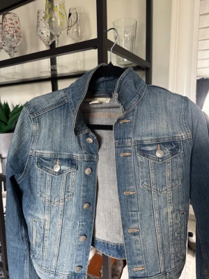 Klassisk blå jeansjacka - Snygg blå jeansjacka med slitna detaljer och silverfärgade knappar. Jackan har två bröstfickor med lock och knapp samt markerade sömmar för en cool look. Perfekt att slänga över en hoodie eller t-shirt för en avslappnad stil.