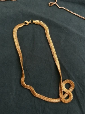 Guldigt halsband i ormkedja 24K - Snyggt halsband i guldton med ormkedjedesign och karbinlås. Märkt med 24K vilket ger en lyxig känsla. Perfekt för dig som gillar stilrena och trendiga accessoarer. Kedjan har en mjuk, böjlig form som ligger snyggt runt halsen.