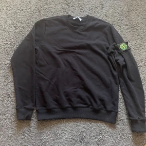 Svart sweatshirt från Stone Island - Svart sweatshirt från Stone Island med klassisk rund halsringning och ribbade muddar. Ikonisk logopatch på vänster ärm som ger en clean och stilren look. Perfekt till jeans eller cargo för en avslappnad streetwearstil. Priset kan diskuteras 