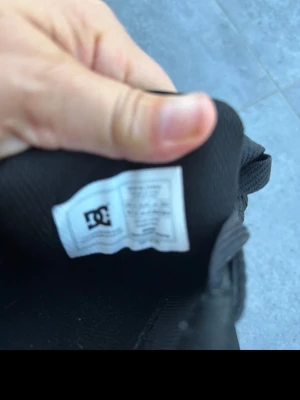 Svarta DC sneakers i mocka - Säljer ett par helsvarta sneakers från DC med klassisk låg profil och tjock sula. Skorna är tillverkade i mocka och har matchande svarta skosnören. Perfekta för dig som gillar streetwear och skate-inspirerad stil.