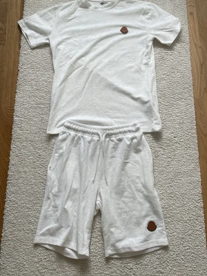 Vitt set med t-shirt och shorts - Säljer ett vitt set med t-shirt och shorts i mjukt material. T-shirten har rund hals och korta ärmar, båda plaggen har en brun patchdetalj. Shortsen har resår i midjan och dragsko för skön passform. Perfekt för chill dagar hemma eller sommarhäng.