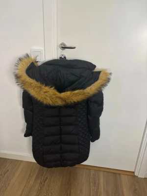 Svart lång dunjacka med fuskpäls - Snygg svart lång dunjacka med huva och fuskpäls i gult runt kanten. Jackan har quiltade detaljer på sidorna och knäppning framtill. Perfekt för kalla vinterdagar och har en modern, figurnära passform.
