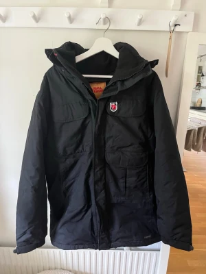 Svart parkas från Fjällräven - Svart parkas från Fjällräven med huva och fyra stora fickor framtill. Jackan har dragkedja och tryckknappar, samt det klassiska Fjällräven-emblemet på bröstet. Perfekt för kalla dagar och tillverkad i slitstarkt material.