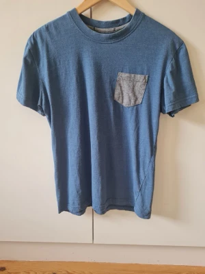 Blå pyjamas t-shirt med grå bröstficka barn - Säljer en enkel blå pyjamas t-shirt med en kontrasterande grå bröstficka. T-shirten har rund halsringning och korta ärmar. För barn. Passar för storlek 158-164 cm.