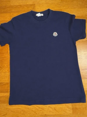 Mörkblå t-shirt från Moncler - Snygg mörkblå t-shirt från Moncler med klassisk passform och rund hals. T-shirten har korta ärmar och ett broderat Moncler-märke på bröstet. Tillverkad i mjuk bomull som känns skön mot huden. Perfekt för en clean och stilren look.