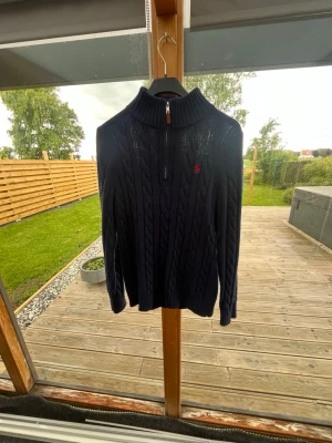 Ramp lauren hoodie  - Snygg mörkblå kabelstickad tröja med halv dragkedja och hög krage. Tröjan har ett diskret rött broderat emblem på bröstet och ribbade muddar vid ärmslut och nederkant. Perfekt för kyliga dagar och enkel att matcha med jeans.