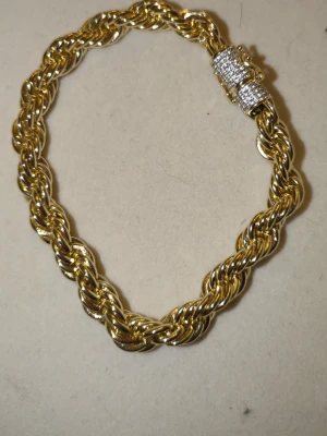 Guldtonat cordell armband 6MM - Snyggt armband i guldfärgad metall med tvinnad kedja och ett glittrande lås dekorerat med små klara stenar. Perfekt accessoar för att lyfta din stil med lite extra bling. Låset är cylinderformat och täckt av gnistrande detaljer.