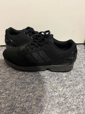 Adidas skor  - Säljer ett par helsvarta Adidas sneakers med klassiska tre ränder på sidan. Skorna har ovandel i meshmaterial och detaljer i plast vid hälen. Snygg och stilren design med snörning och bekväm sula. Perfekta för dig som gillar sportig och clean stil.
