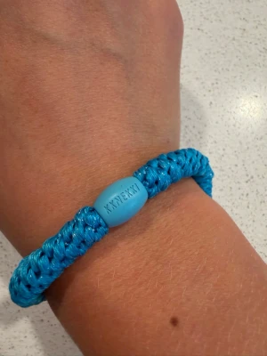 Blått armband från Kknekki - Snyggt blått armband från Kknekki med flätad design i glansigt syntetmaterial. Armbandet har en oval pärldetalj med Kknekki-loggan ingraverad. Perfekt accessoar för att addera färg till din stil.