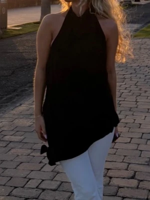Svart halterneck topp med öppen rygg - Säljer en svart halterneck topp med asymmetrisk nederkant och helt öppen rygg. Toppen är ärmlös och har en lös passform, perfekt för varma dagar. Materialet ser ut att vara mjukt och följsamt, troligen polyester eller liknande.