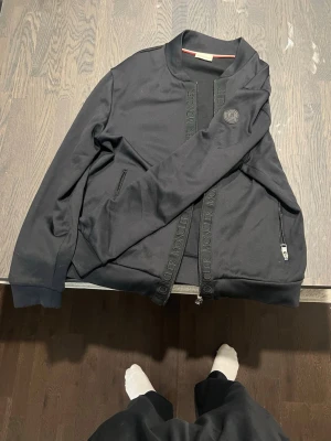 Svart zip hoodie från Moncler - Snygg svart hoodie från Moncler med dragkedja framtill och diskret logga på bröstet. Hoodien har två fickor med dragkedja och ribbade muddar. Tillverkad i mjukt bomullsmaterial och har Moncler-text längs dragkedjan.