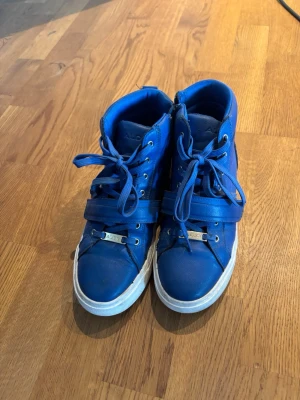Blå höga sneakers med snörning - Snygga blå sneakers i high-top modell med vita sulor och snörning. Skorna har även ett band med metallspänne över vristen för extra stil. Perfekta för dig som vill sticka ut med färg på fötterna.