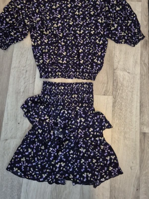 Blommigt set i mörkblått och lila - Säljer ett tvådelat set med topp och kjol i mörkblått tyg med små blommor i lila, vitt och gult. Toppen har korta puffärmar och resår i midjan, kjolen har volanger och smock i midjan. Perfekt för dig som gillar söta och trendiga plagg med blommigt mönster.