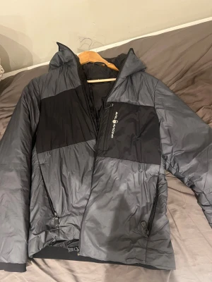Grå och svart pufferjacka All Racing - Snygg pufferjacka i grått och svart från All Racing. Jackan har huva, dragkedja framtill och två sidofickor med dragkedja. Tillverkad i ett glansigt, vadderat material som håller värmen under kalla dagar. Perfekt för vintern och riktigt stilren look.