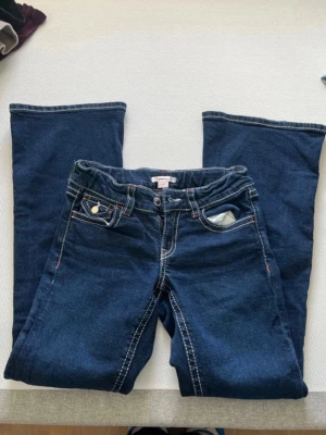 Mörkblå bootcut jeans med kontrastsömmar - Snygga mörkblå bootcut jeans med vita kontrastsömmar och dekorativa fickor både fram och bak. Jeansen har knappdetaljer på fickorna och resår i midjan bak för extra komfort. Perfekta för dig som gillar en klassisk men trendig look.