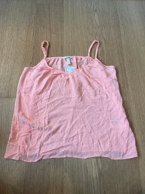 Ljusrosa linnetopp från H&M - Säljer en ljusrosa linnetopp från H&M med tunna axelband och v-ringad hals. Toppen är enkel och luftig, perfekt för varma dagar. Materialet är mjukt och lätt, vilket gör den skön att bära.