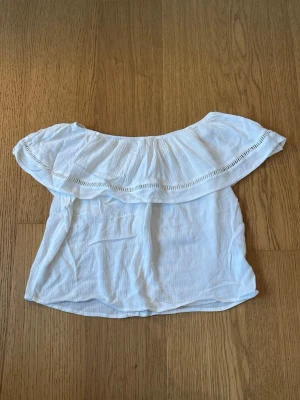Vit offshoulder blus med volang - Säljer en söt vit offshoulder blus med bred volang runt axlarna och fina håldetaljer. Toppen har en luftig och lätt känsla, perfekt för varma dagar. Knytning i ryggen för extra detalj. Passar dig som gillar en romantisk och avslappnad stil.