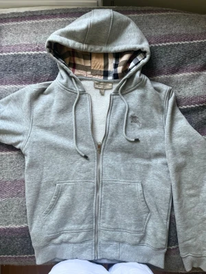 Burberry zip, grå - Snygg grå hoodie från Burberry med dragkedja framtill och klassiskt rutigt foder i huvan. Hoodien har två fickor fram och justerbara snören i huvan. Har en skön passform. Strl M passar både S och M nog