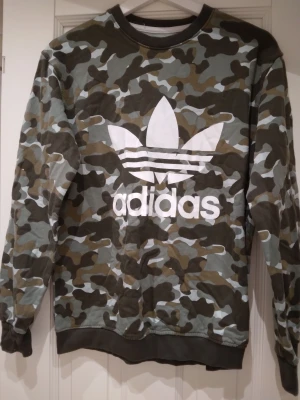 Adidas camo sweatshirt med logga, camouflage, grön, Small - Säljer en camouflagemönstrad sweatshirt från Adidas med stor vit klassisk Adidas logga och text på bröstet. Tröjan har rund hals, långa ärmar och ribbade muddar vid ärmslut och nederkant. Perfekt för en streetwear-look. Mycket bra skick. Strlk Small.