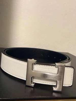 Vitt skärp med stor H-spänne - Snyggt vitt bälte med en stor, silverfärgad H-formad metallspänne. Bältet har svart insida och är gjort i slätt och präglat skinn. Perfekt accessoar för att lyfta din outfit.