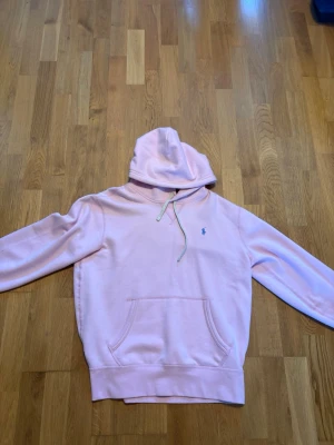 Ljusrosa hoodie från Polo Ralph Lauren - Snygg ljusrosa hoodie med huva och dragsko från Polo Ralph Lauren. Klassisk känguruficka framtill och broderad logga på bröstet. Perfekt för en avslappnad och trendig stil.