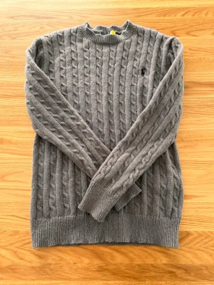 Grå kabelstickad tröja - Kabelstickad grå tröja med rund hals och långa ärmar. Tröjan har ribbstickade muddar vid ärmslut och nederkant. 