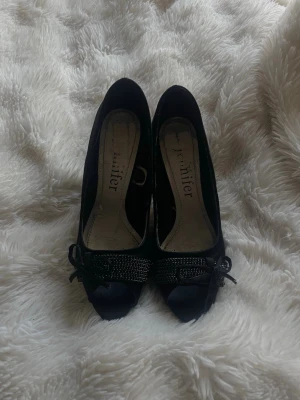 Svarta vintage pumps med rosett - Snygga svarta vintage pumps med öppen tå och dekorativ rosett framtill. Skorna har en glittrig detalj över tån och smal klack. Materialet ser ut att vara satinliknande tyg som ger en elegant känsla. Perfekta för dig som vill ha något extra till outfiten.