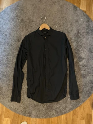 Svart långärmad skjorta - Stilren svart skjorta med klassisk krage och knappar framtill. Skjortan är långärmad och har en rak passform. Tillverkad i mjuk bomull som känns skön mot huden. Perfekt för dig som gillar en clean och enkel look.