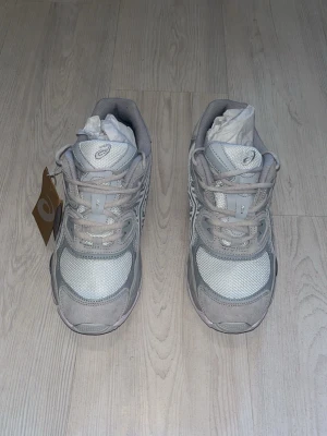 Gråvita sneakers från Asics - Snygga sneakers från Asics i grått och vitt med mesh och mockadetaljer. Klassisk låg modell med snörning och bekväm sula. Perfekta för dig som gillar stilrena och sportiga skor.
