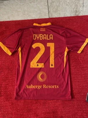 Röd AS Roma Dybala matchtröja - Säljer en röd AS Roma matchtröja med gula detaljer, Dybala och nummer 21 på ryggen. Framsidan har SPQR-tryck och vargens symbol. Tillverkad i lätt och ventilerande material, perfekt för fotboll eller träning.