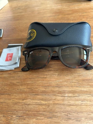 Ray-Ban Wayfarer solglasögon - Säljer ett par klassiska Ray-Ban Wayfarer solglasögon i brunmelerad plast med mörka glas. Medföljer originalfodral i svart skinn och putsduk. Perfekt accessoar för soliga dagar och ikonisk look.