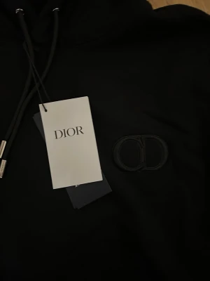 Svart hoodie från Dior med logga - Snygg svart hoodie från Dior med broderad CD-logga på bröstet. Klassisk modell med huva, dragsko och magficka. Tillverkad i mjukt material som känns lyxigt och bekvämt. Perfekt för dig som vill ha en stilren och exklusiv look.
