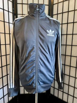 Adidas Originals Grå Track Jacket M – Big Trefoil Retro Y2K - Riktigt snygg Adidas Originals track jacket i mörkgrått med ikoniska vita 3-Stripes längs ärmarna. Stor broderad outline-Trefoil över ryggen som verkligen lyfter hela jackan, plus broderad Trefoil på bröstet och loggad zip. Clean retro/Y2K-look med hög krage och smal klassisk passform. Märkt M. Pit to pit 51 cm, axel 45 cm, längd 66 cm. 100% polyester, Made in Turkey. Fint begagnat skick – en riktig statement-piece! 