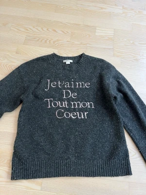 Mörkgrå stickad tröja med text - Mysig mörkgrå stickad tröja med rund hals och broderad text i vitt och rosa på framsidan: 'Je t’aime de tout mon coeur'. Tröjan har långa ärmar och ribbade muddar vid ärmslut och nederkant. Perfekt för kalla dagar.
