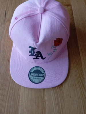 Rosa LA keps - Säljer en rosa snapback keps med broderad LA-logga i svart och en röd ros på framsidan. Kepsen har platt skärm och justerbar snapback baktill. Materialet är bomullstyg och designen är riktigt streetinspirerad.