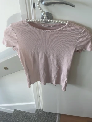 Ljusrosa ribbad croppad topp - Söt ljusrosa croppad topp med ribbad struktur och korta ärmar. Toppen har vågig kant nertill och vid ärmsluten för en extra detalj. Perfekt till jeans eller kjol för en trendig look.
