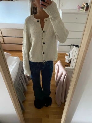 Vit fluffig kofta från Vero Moda - Mysig vit kofta från Vero Moda med fluffig textur och svarta knappar framtill. Koftan har lång ärm, rund halsringning och en avslappnad passform. 