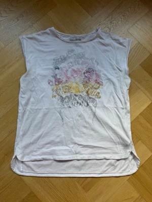 Vit ärmlös topp Zadig & Voltaire - Säljer en vit ärmlös topp från Zadig & Voltaire med ett blommigt tryck i rosa, gult och grått framtill. Toppen har rund halsringning och är tillverkad i mjuk bomull, perfekt för en avslappnad och trendig look. Strl 12 år/xs