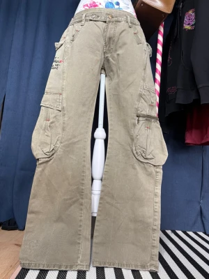 Y2K cargo byxor khaki med broderier & stora fickor - Snygga Y2K-inspirerade cargobyxor i en tvättad khakigrön färg med breda ben och flera stora cargofickor. Byxorna har röda kontrastsömmar, broderade textdetaljer och en riktigt cool utility-look som passar perfekt till grunge, streetwear och alternative outfits.  • Storlek: 44 • Midjemått: 88 cm • Innerbenslängd: 80 cm • Färg: Khaki/olivgrön • Material: 100% bomull • Bra vintage-skick.